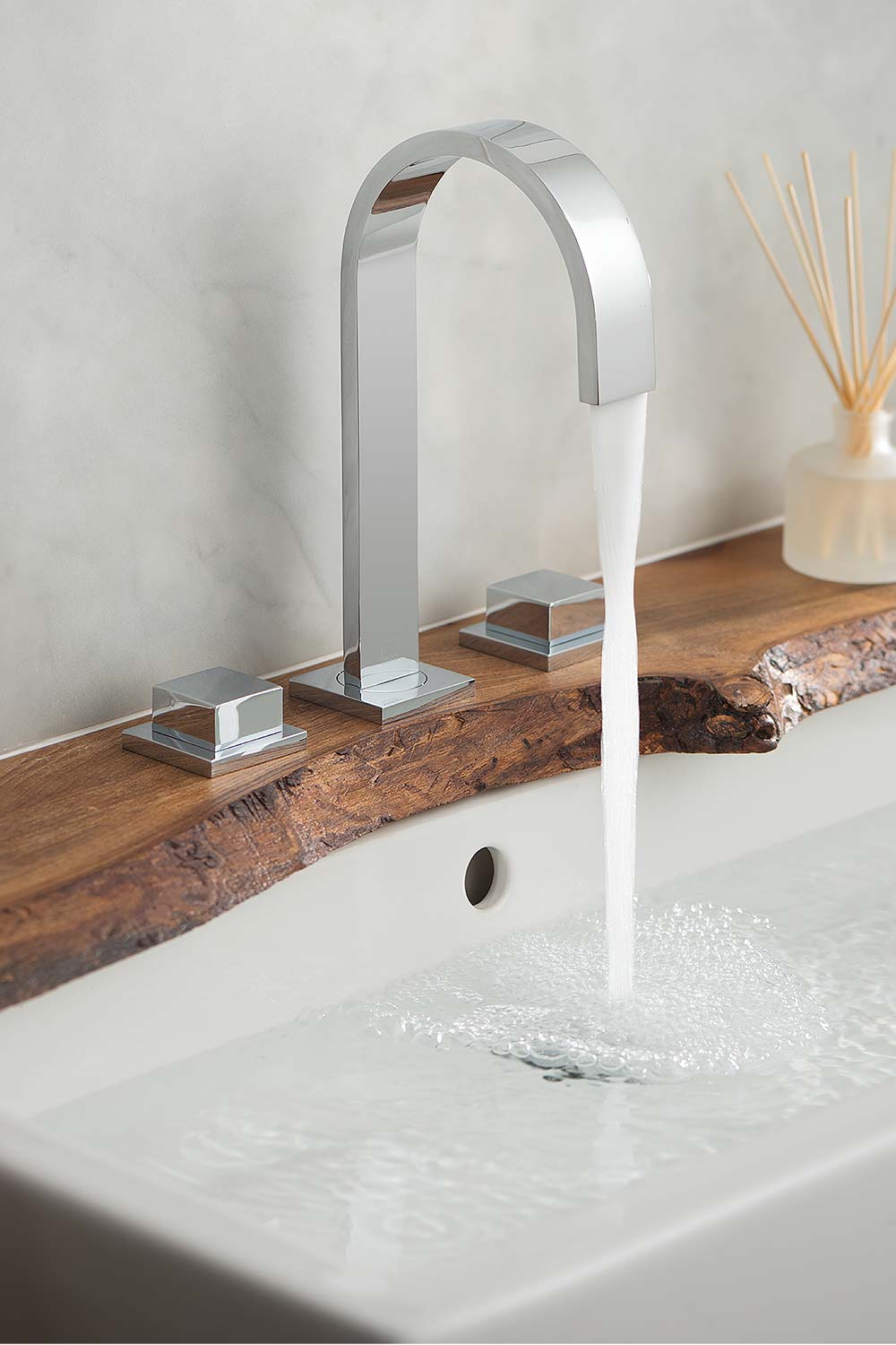 Modern three hole basin mixer with square handles and water flowing from the tall spout on a rough edge elm wood shelf over a white basin.