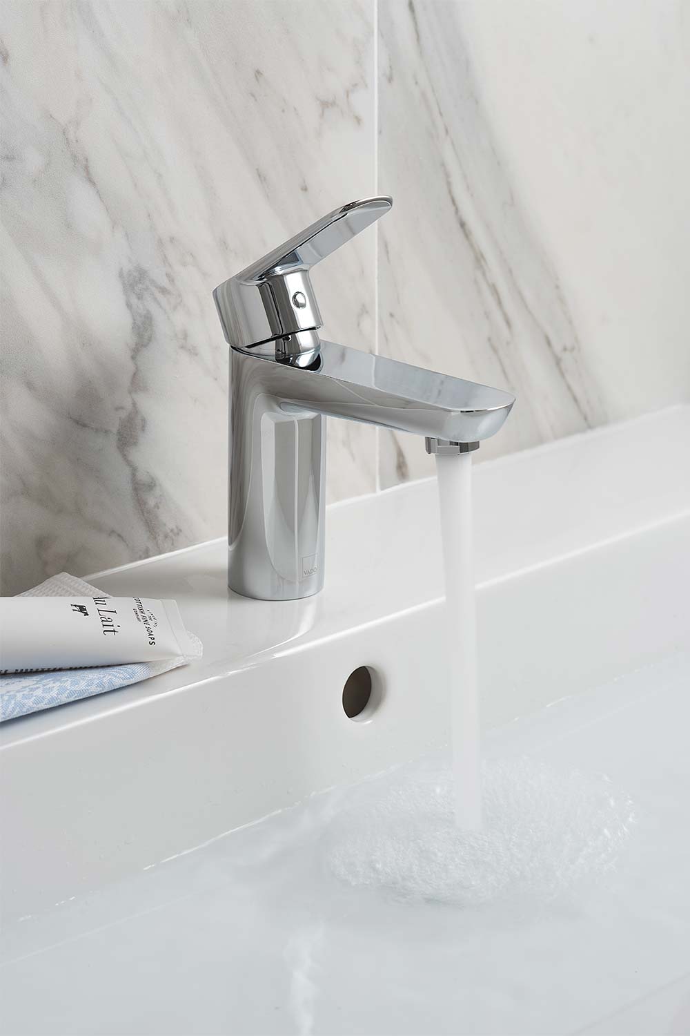 Single lever mono basin mixer with water flowing from the spout on a white basin with a marble tiled wall.