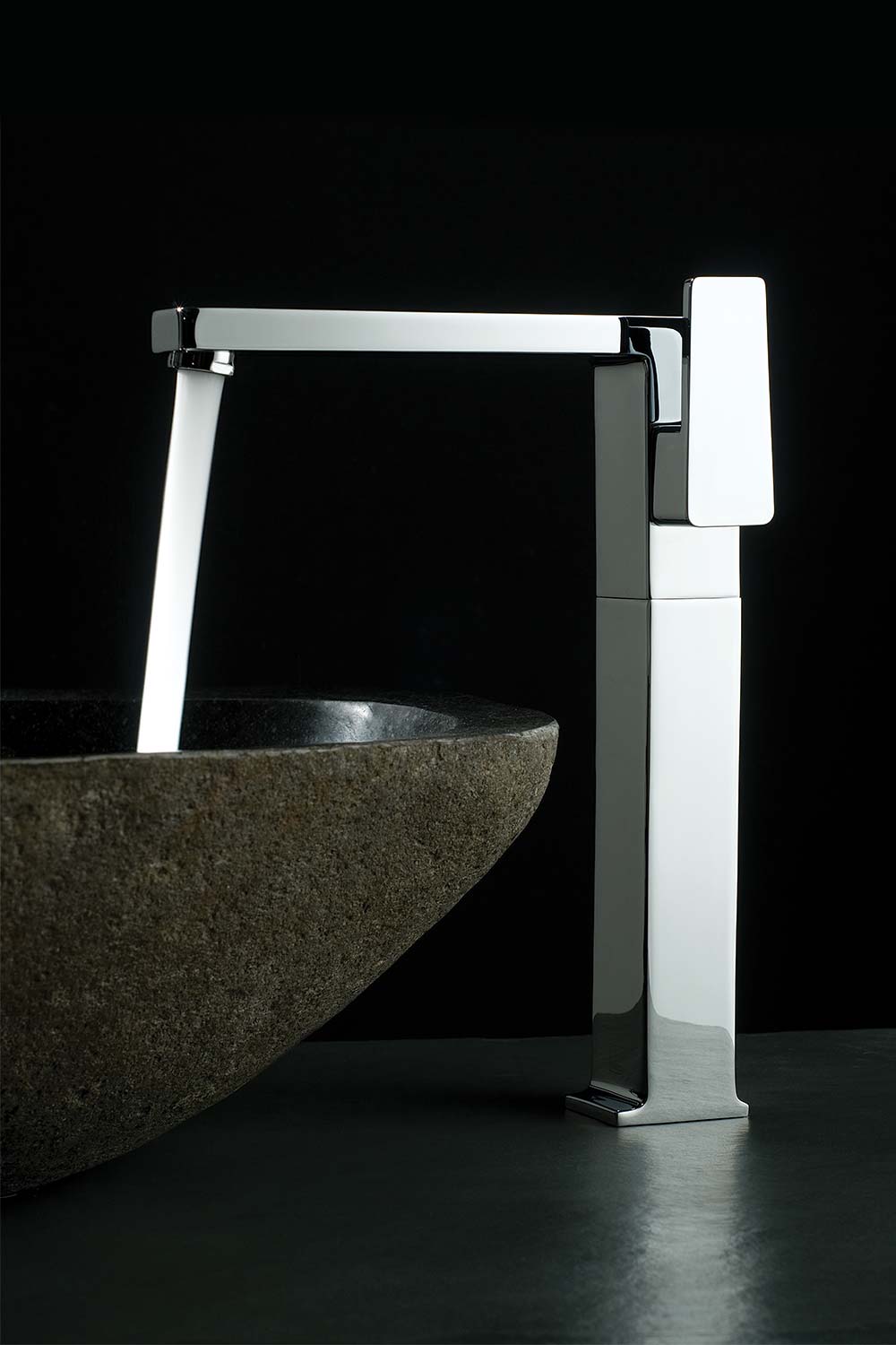 Modern single lever extended mono basin mixer over a stone basin against a black background.