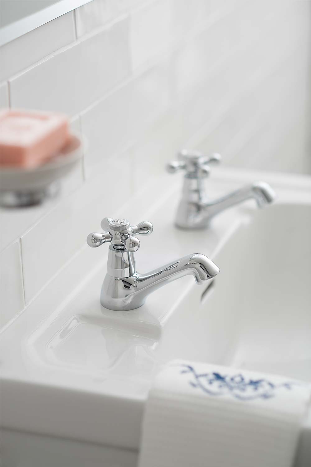 A pair of traditional basin pillar taps on a basin with white tiles, a hand towel and soap in a dish.