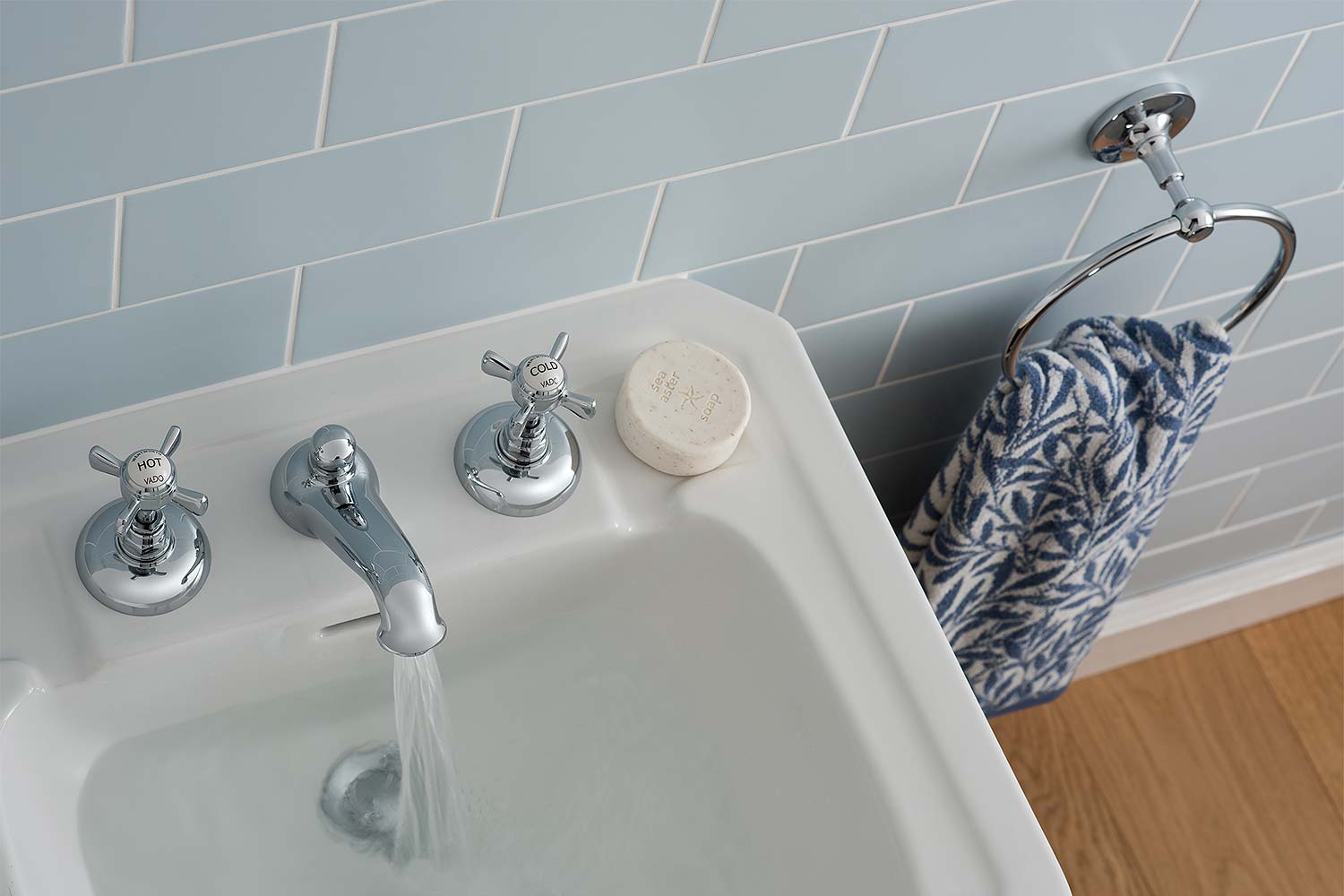 A traditional three hole deck mounted basin mixer on a white basin with a light blue tiled wall, and towel ring with towel.