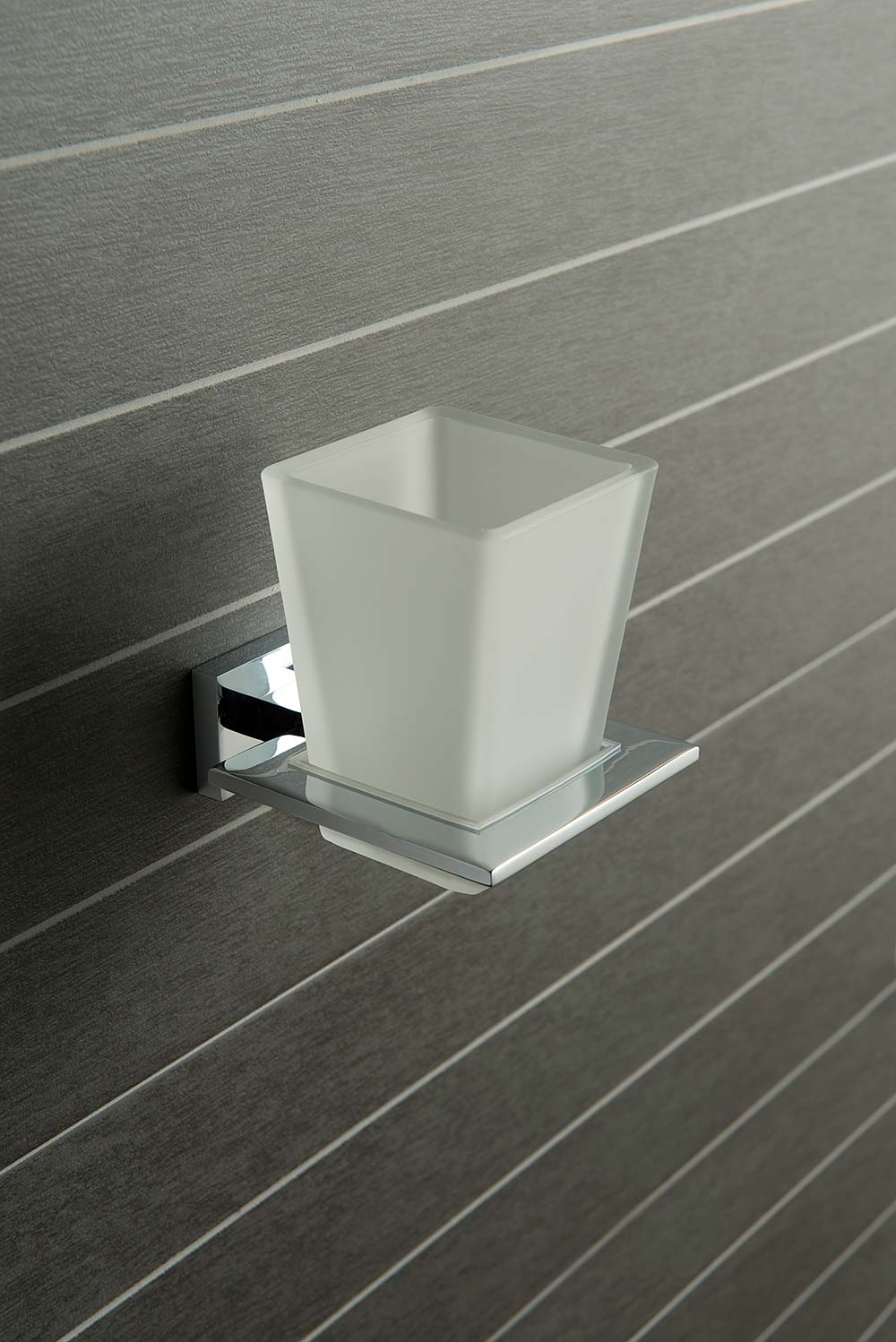 A modern chrome square tumbler and holder on a linear pattern grey tiled wall.
