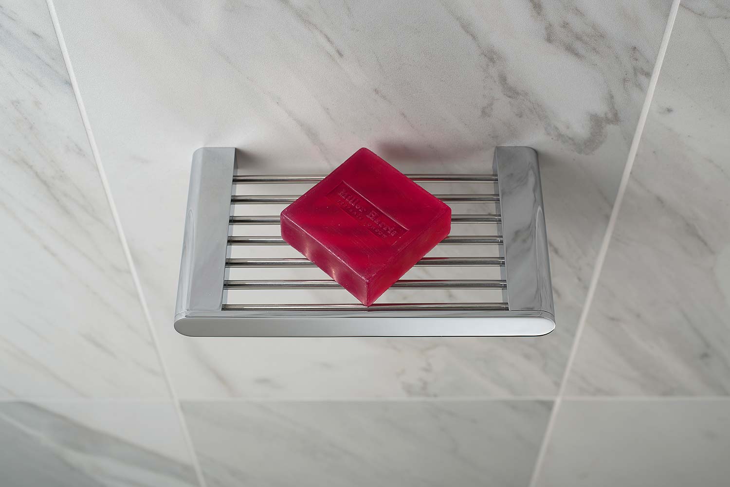 A bright red square soap on a modern chrome soap dish on a marble tiled wall.