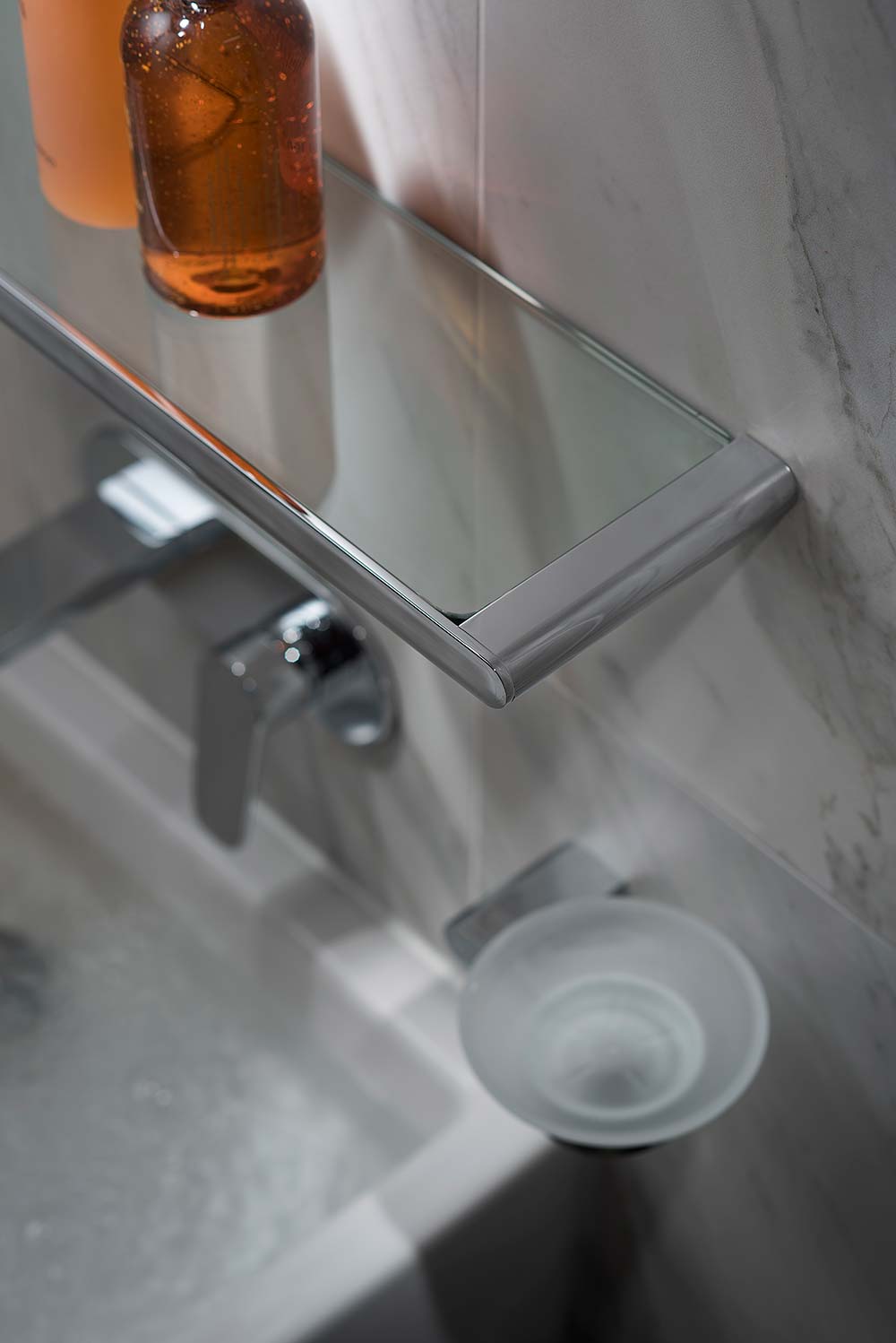 Modern chrome and glass shelf on a marble tiled wall above a basin mixer and basin.