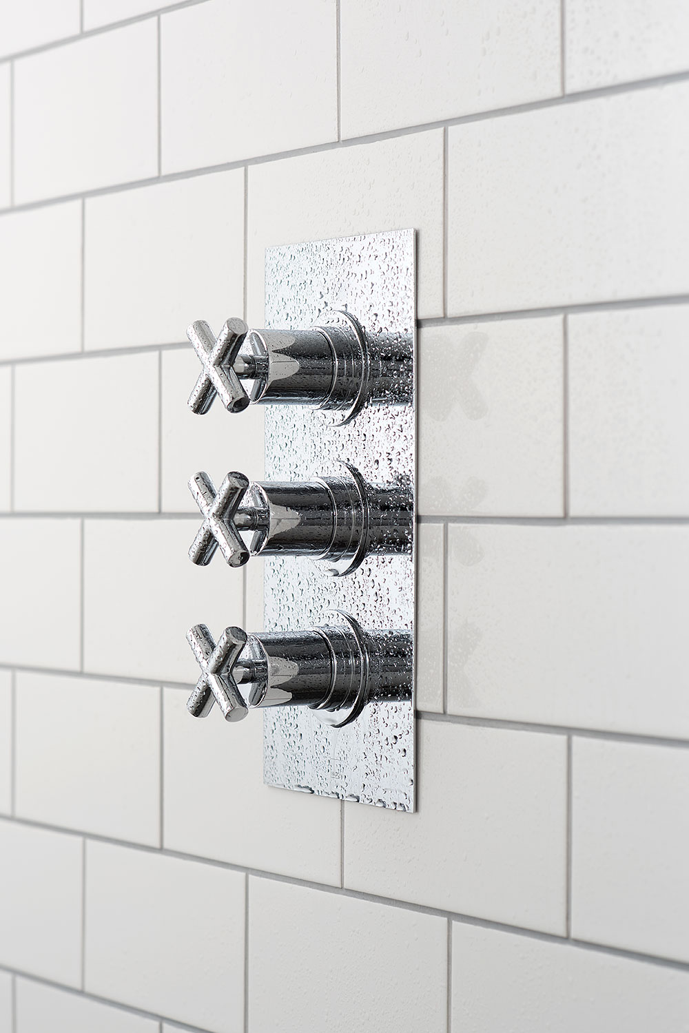 Side view of a three handle thermostatic shower valve on a white tiled wall with grey grout.