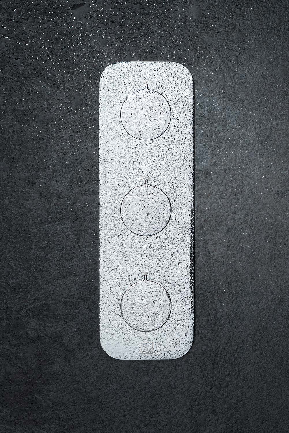 Square on photograph of a three handle shower valve covered in water drops on a dark grey wall