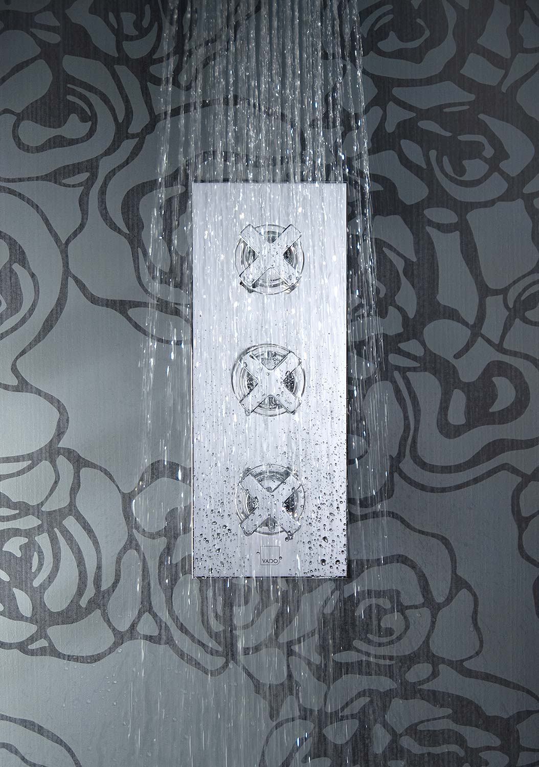 Square on photograph of a three handle shower valve with water falling in front of it on a graphic grey tiled wall.