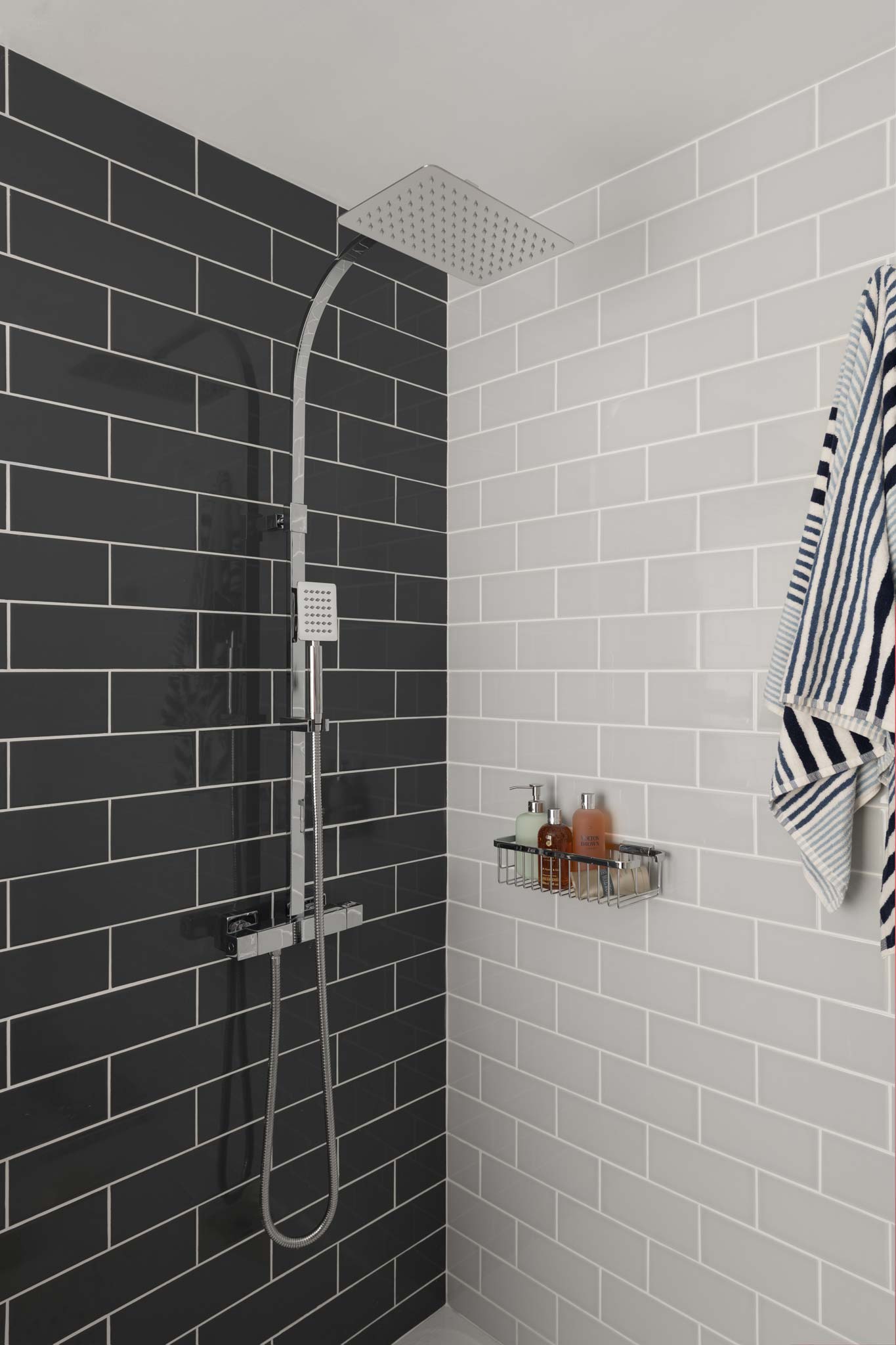 A wall mounted thermostatic shower mixer with a rigid riser on a tiled wall with a basket of shower bottles and a towel