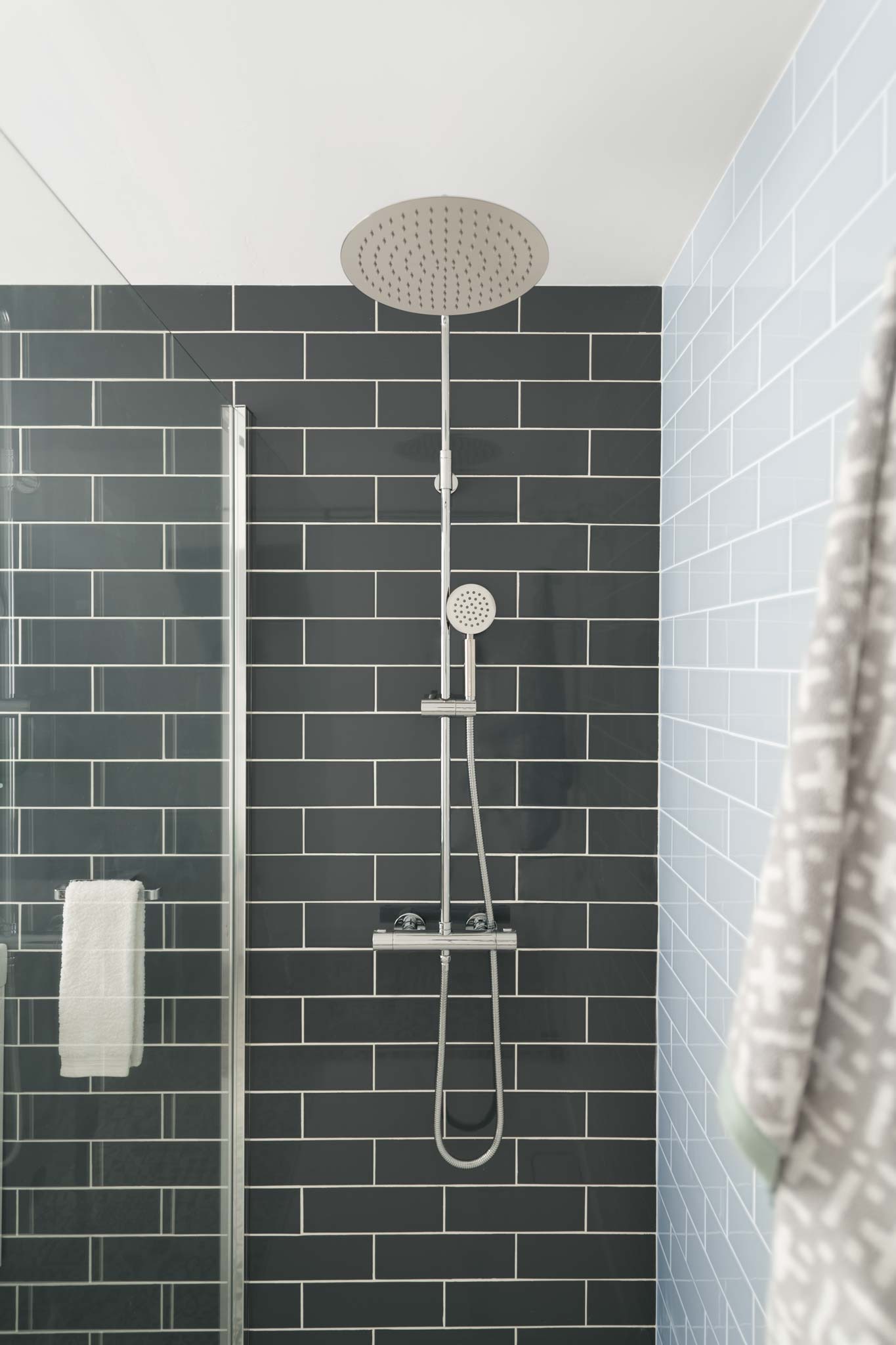 A wall mounted thermostatic shower mixer with a rigid riser on a tiled wall with a shower screen and towels