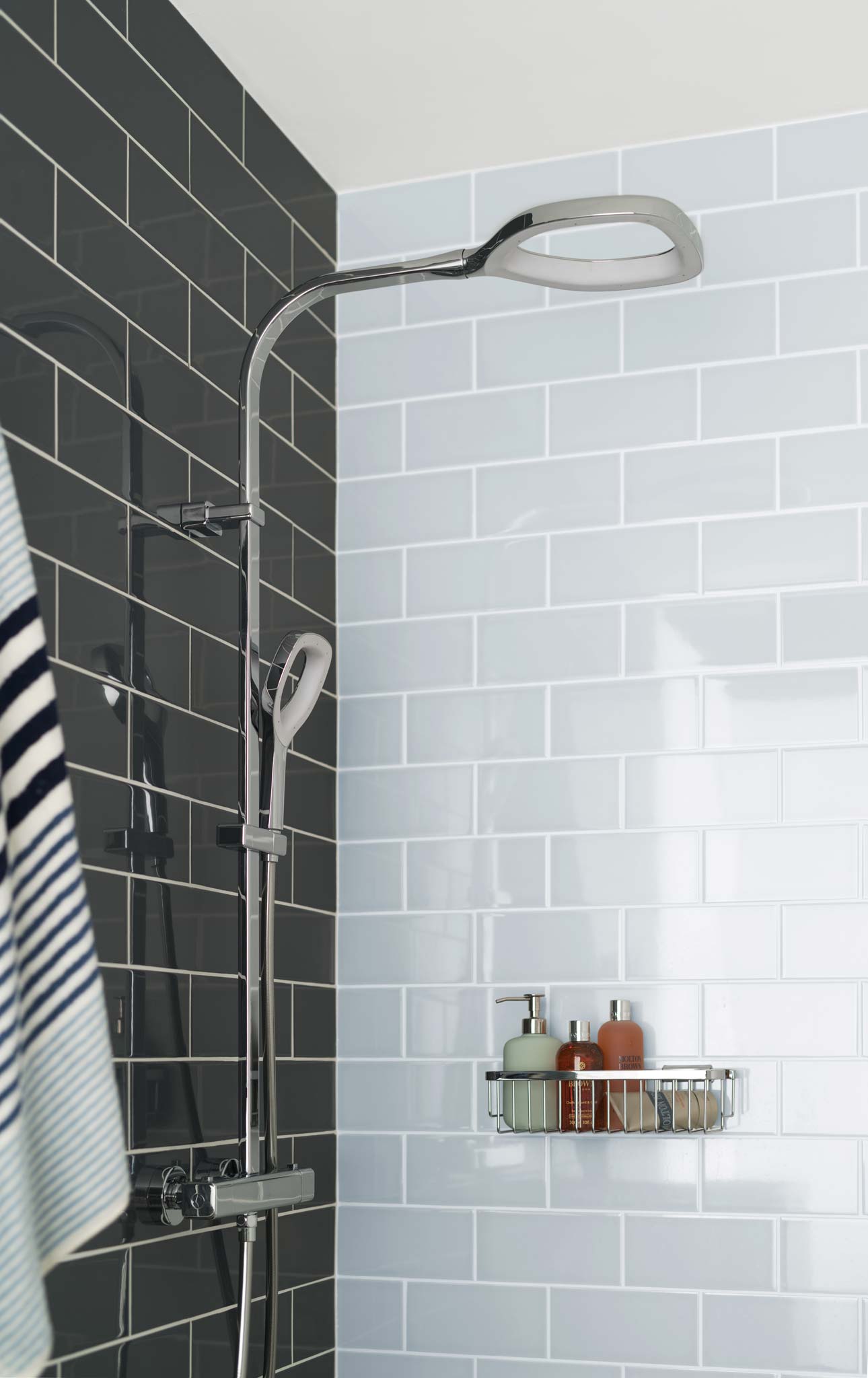 A wall mounted thermostatic shower mixer with a rigid riser on a tiled wall with a basket of shower bottles and a towel