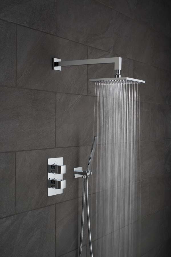 Modern square and rectangular shower head and arm, wall mounted shower kit and concealed thermostatic shower valve on a black slate tiled wall with water cascading out of the shower head