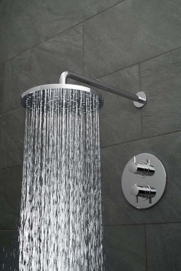 Round shower head and arm with a round concealed thermostatic shower valve on a black slate tiled wall, with water cascading out of the shower head.