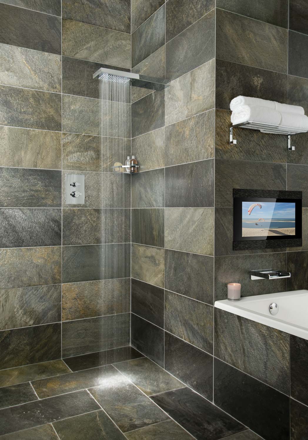 Tiled bathroom with a bath, shower, accessory basket, rectangular wall mounted thermostatic valve and water cascading from the shower head